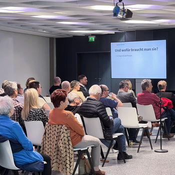 viele Menschen sitzen in Stuhlreihen und hören einem Vortrag zu
