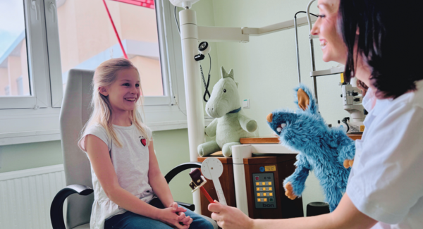 Kind sitzt lachend auf Untersuchungsstuhl, Ärztin hält Kuscheltier in der Hand