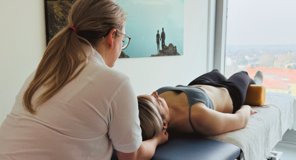 Patientin liegt auf Liege, Physiotherapeutin umfasst ihren Nacken