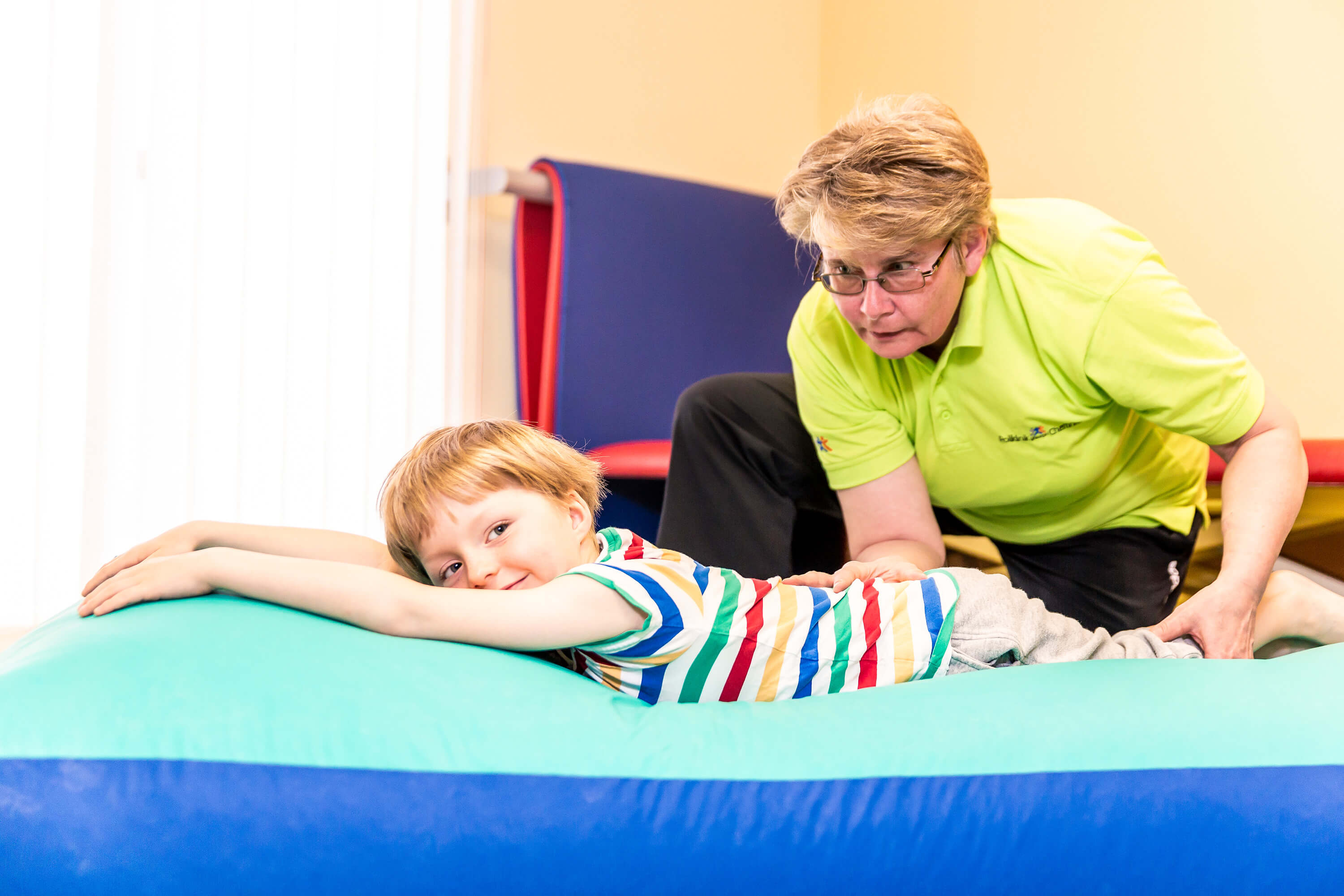 Kind liegt auf Matte, Physiotherapeutin korrigiert Haltung